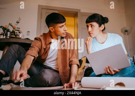 Männlicher Freelancer, der mit einer Kollegin spricht, die einen Laptop hält, während er im Home Office sitzt Stockfoto