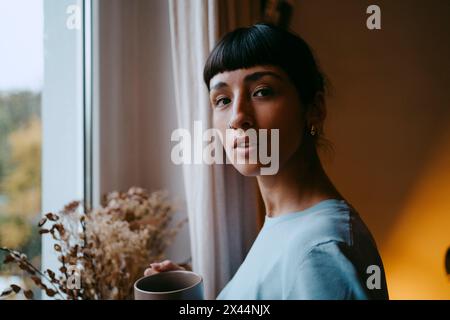 Seitenporträt einer jungen Frau, die zu Hause eine Kaffeetasse in der Nähe eines Fensters hält Stockfoto