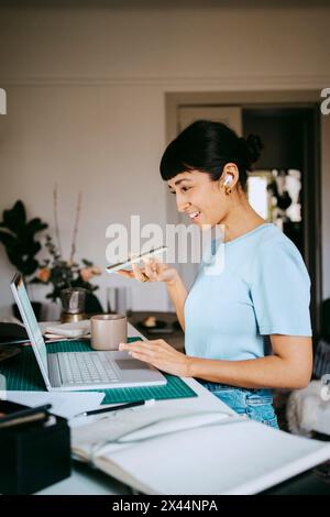 Lächelnde junge Freiberuflerin, die über Lautsprechertelefon spricht, während sie im Heimbüro einen Laptop benutzt Stockfoto