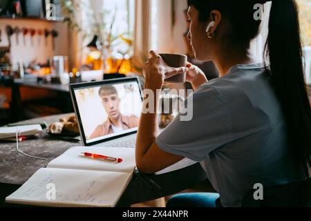 Junge Freiberuflerin, die eine Kaffeetasse hält, während sie im Heimbüro Videogespräche mit einem Kollegen auf einem Laptop führt Stockfoto