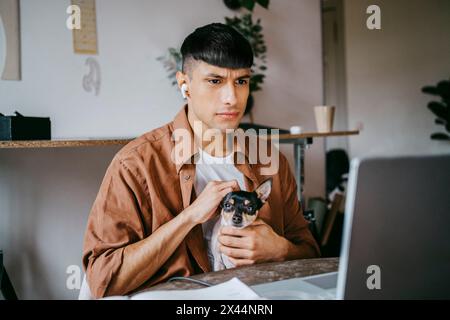Ernster männlicher Freelancer streichelt Hund, während er im Home Office auf den Laptop blickt Stockfoto