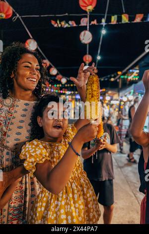 Ein aufgeregtes Mädchen, das gegrilltes Getreide hält, während er im Urlaub neben der Mutter auf dem Lebensmittelmarkt steht Stockfoto