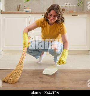 Eine Frau fegt den Boden mit einem Besen und einer Schmutzpfanne in ihrer Küche. Weibliche Hände in gelben Handschuhen beim Reinigen der Küche Stockfoto