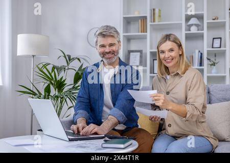 Ein reifes Paar sitzt bequem auf der Couch und lächelt, während sie ihre Finanzen gemeinsam verwalten. Die Szene ist gemütlich, mit einer friedlichen Kulisse für das Home Office. Stockfoto