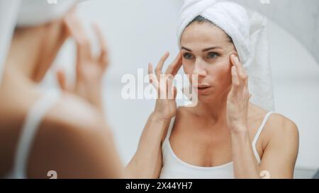 Die schöne kaukasische Frau berührt ihr perfektes Gesicht, genießt ihre ideale faltenfreie Haut und lächelt sinnlich in den Badezimmerspiegel. Happy Middle Age Female Verwendet Natürliche Kosmetische Hautpflegeprodukte Stockfoto