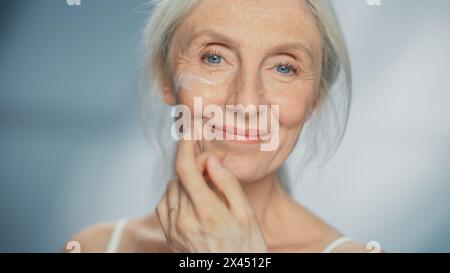 Porträt der schönen älteren Frau, die sanft Gesichtscreme aufträgt. Ältere Dame macht ihre Haut weich, glatt, knitterfrei mit natürlicher Anti-Aging-Kosmetik. Produkt für Beauty Hautpflege, Make-up Stockfoto