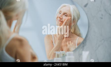 Schöne Seniorin schaut ins Badezimmer Spiegel berührt ihr Gesicht, lächelt glücklich, genießt ihr Aussehen und die morgendliche Routine. Würde und Gnade im Alter. Wohlbefinden, Hautpflege, Schönheitsprodukte Stockfoto