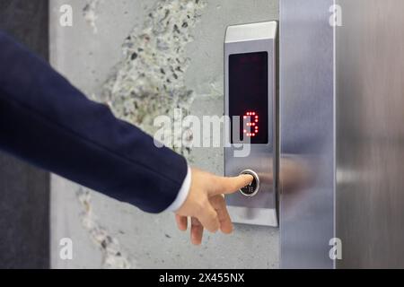 Ein gut gekleideter Geschäftsmann drückt einen Fahrstuhlknopf und signalisiert die dritte Etage eines modernen Bürogebäudes. Das Bild erfasst Technologie und Zugänglichkeit in der Unternehmenswelt. Stockfoto