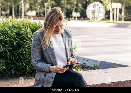 Blonde Geschäftsfrau im Anzug, die draußen ein Tablet benutzt und lächelt Stockfoto
