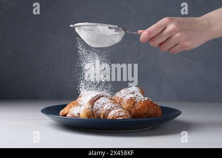 Frau mit einem Sieb, das Puderzucker auf Croissants streut, am weißen Holztisch, Nahaufnahme Stockfoto