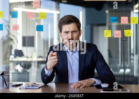 Ein verärgerter Geschäftsmann, der Frustration ausdrückt und zeigt, während er während eines Videoanrufs in einer modernen Büroumgebung eine hitzige Diskussion führt. Stockfoto