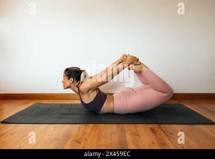 Junge mittelgroße Frau in einem rosa Sportoutfit, die zu Hause Yoga auf einer Matte macht. Bogenstellung. BRUSTÖFFNENDE YOGA-POSEN Stockfoto