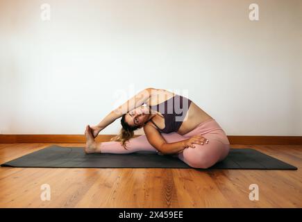 Junge mittelgroße Frau in einem rosa Sportoutfit, die zu Hause Yoga auf einer Matte macht. Gedrehte Kopf-Knie-Haltung Stockfoto