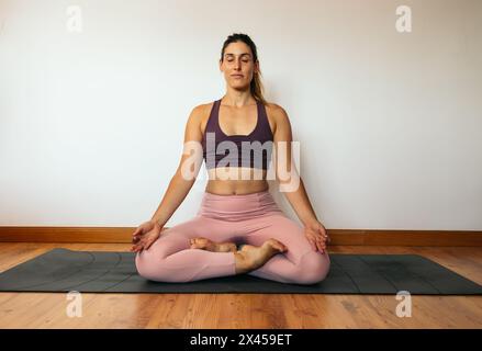 Junge mittelgroße Frau in einem rosa Sportoutfit, die zu Hause Yoga auf einer Matte macht. Lotus Pose Stockfoto