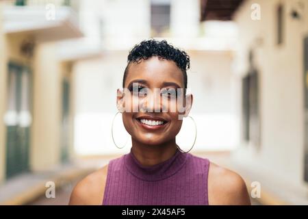 Porträt einer stilvollen Frau mit kurzen Haaren und großen Ohrringen, die die Kamera lächelnd ansieht. Sie sieht selbstbewusst aus und gibt gute Vibes ab Stockfoto