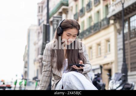 Asiatische junge Frau, die in einer europäischen Stadt auf einer Bank sitzt und auf jemanden wartet, während sie auf das Smartphone blickt und Musik mit Kopfhörern hört Stockfoto