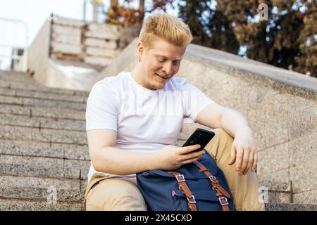 Albino latin Mann sitzt auf einer Treppe auf der Straße mit Smartphone. Er trägt weiße Kleider und lächelt auf das Handy Stockfoto