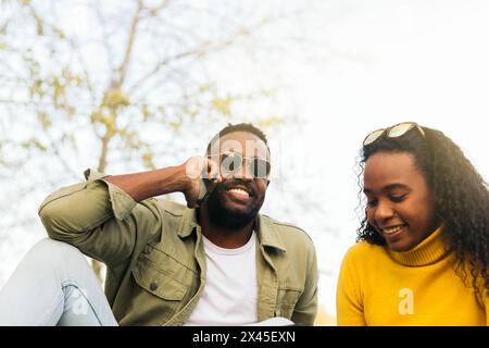 Lächelndes afro amerikanisches Paar, das auf dem Gras mit dem Handy sitzt Und telefonieren Stockfoto