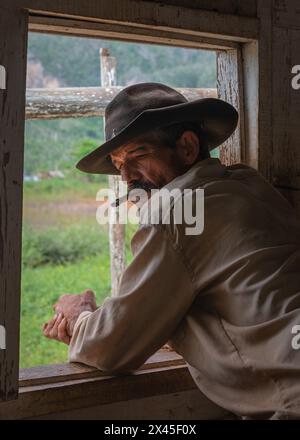 Ein Tabakbauer mit Cowboyhut raucht eine Zigarre am Fenster seines Bauernhauses im Vinales Valley, Vinales, Kuba. Stockfoto