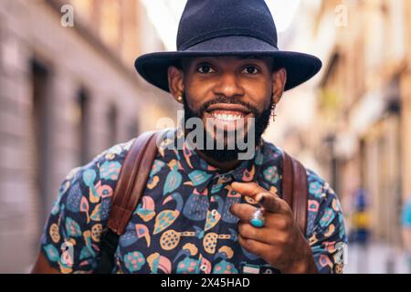 Stilvoller schwarzer Mann mit alternativem Hipster-Look, der an einem sonnigen Sommertag durch die Straßen einer Stadt spaziert. Er lächelt glücklich Stockfoto