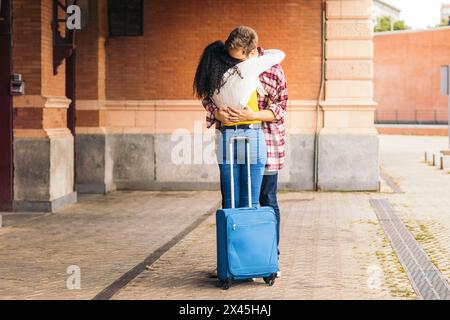 Multirassisches junges erwachsenes Paar, das sich umarmt und küsst, nachdem er sich wieder auf dem Bahnhof getroffen hat. Sie trägt einen blauen Koffer von ihrer Reise und er wartet darauf Stockfoto