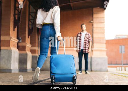Multirassisches junges erwachsenes Paar, das sich umarmt und küsst, nachdem er sich wieder auf dem Bahnhof getroffen hat. Sie trägt einen blauen Koffer von ihrer Reise und er wartet darauf Stockfoto