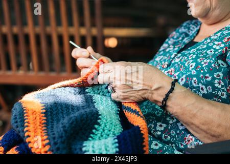 Eine ältere Frau, die eine Decke häkelt. Sie konzentriert sich auf ihre Aufgabe, sitzt auf einem Stuhl auf der Terrasse ihres Hauses. Stockfoto