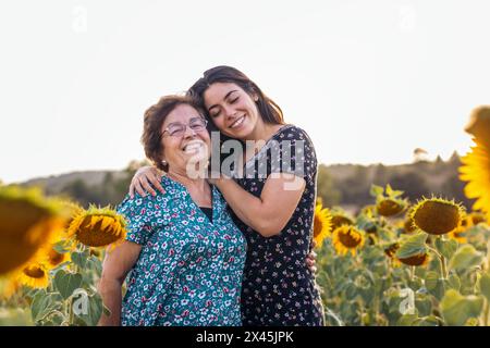 Lächelnde Seniorin in einem wunderschönen Sonnenblumenfeld bei Sonnenuntergang mit ihrer Enkelin, die die gemeinsame Zeit genießt Stockfoto