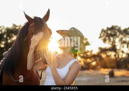 Junge Frau mit Hut, während sie mit ihrem Pferd auf dem Land spaziert, während die Sonne hinter ihr untergeht Stockfoto