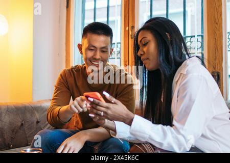 Junger asiatischer Mann und lateinische Frau, die auf dem Sofa sitzen und sich entspannt mit dem Smartphone unterhalten. Sie zeigt ihm ihre sozialen Medien auf dem Bildschirm Stockfoto