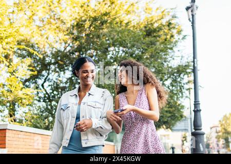 Zwei lateinamerikanische Freunde laufen die Straße entlang, haben Spaß und lachen, während sie ein Smartphone benutzen. Stockfoto
