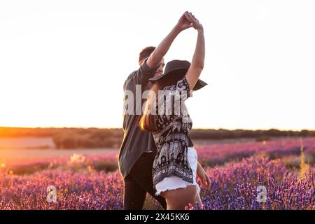 Wunderschönes hintergrundbeleuchtetes Foto eines jungen, liebevollen Paares, das bei Sonnenuntergang in einem Lavendelfeld tanzt. Stockfoto