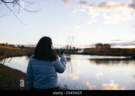 Rückansicht einer jungen Frau im Daunenmantel, die den Sonnenuntergang auf einem See in der Natur beobachtet und Fotos mit dem Smartphone macht Stockfoto