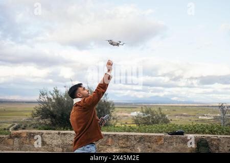 Junger Mann, der eine Drohne benutzt, um die Landschaft aufzunehmen und zu fotografieren. Er hält den Controller in der einen Hand und mit der anderen wird er die Drohne fangen Stockfoto