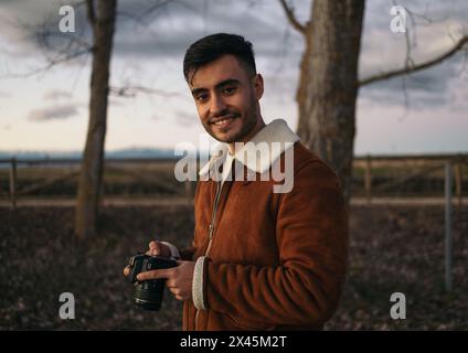 Porträt eines lächelnden Fotografen, der in die Kamera schaut. Junger Mann in brauner Jacke, der sich die Fotos auf der Kamera ansieht Stockfoto