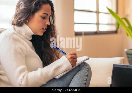 Eine junge Frau, die zu Hause sitzt und mit ihrem Laptop und einem Notizbuch für die Universität studiert. Der Teilnehmer macht sich Notizen, während er sich die Informationen online ansieht Stockfoto