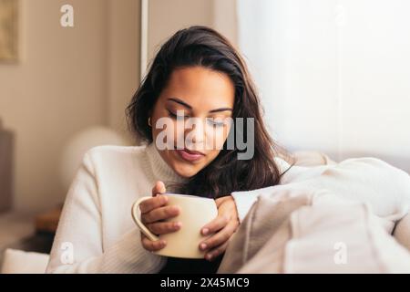 Eine junge Frau genießt eine Tasse Kaffee, während sie auf dem Sofa in der Nähe eines Fensters in ihrem Haus sitzt. Stockfoto