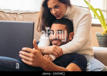 Ein gemütliches und romantisches Stockfoto eines jungen Paares, das zu Hause auf einer Couch sitzt und sich einen Film auf einem Laptop ansieht. Stockfoto