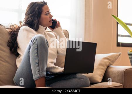 Eine junge Frau, die auf ihrer Heimcouch sitzt und an ihrem Laptop arbeitet, während sie mit einem Smartphone telefoniert. Stockfoto