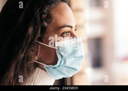 Eine schöne junge Frau, die während der COVID-19-Pandemie aus dem Fenster in ihrem Haus blickt. Stockfoto