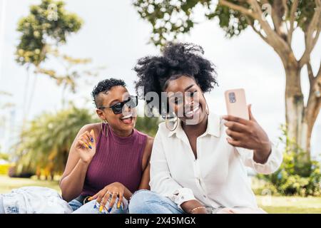 Zwei schwarze Freundinnen, die Spaß haben und Selfies mit dem Smartphone machen, sitzen auf dem Gras im Park Stockfoto