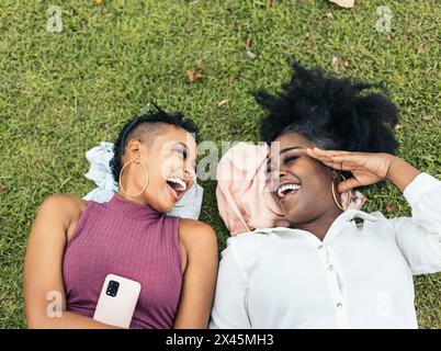 Zwei schwarze Freundinnen, die Spaß haben und ein Smartphone benutzen, das auf dem Gras im Park liegt Stockfoto