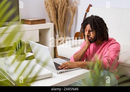 Besorgter und depressiver junger Mann, der von zu Hause aus arbeitet und sein Gesicht mit den Händen vor seinem Laptop bedeckt und schlechte Nachrichten erhält Stockfoto