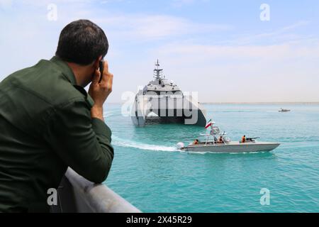 Persischer Golf, Buschehr, Iran. April 2024. Ein Mitglied des Korps der Islamischen Revolutionsgarde (IRGC) spricht mit seinem Handy, während er das Kriegsschiff der Islamischen Revolutionsgarde (IRGCN) Shahid (Martyr) Hassan Bagheri und ein Schnellboot der Islamischen Revolutionsgarde (IRGC) ansieht. die während der Marineparade der IRGC zum Nationalfeiertag des Persischen Golfs in der Nähe des Kernkraftwerks Bushehr in der Seehafenstadt Bushehr, Provinz Bushehr, im Süden des Iran, entlang des Persischen Golfs segeln. Der Iran feiert den Jahrestag der Befreiung des Südens des Landes aus Stockfoto