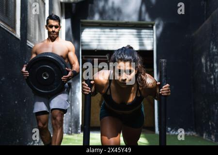 Starke hispanische Frau in Sportkleidung, die während des intensiven Powerlifting-Trainings auf der Straße in der Nähe des Gebäudes einen schweren Schlitten mit Gewichten in die Nähe des Trainers schiebt Stockfoto