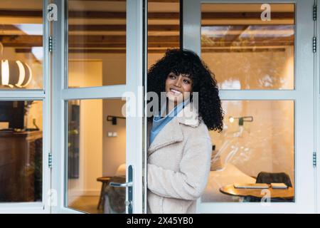 Fröhliche junge afroamerikanische Frau mit lockigen dunklen Haaren in warmem Mantel, die durch eine Glastür ins Haus kommt und wegschaut Stockfoto