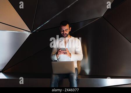 Fokussierter bärtiger Mann mit Netbook-Textnachrichten auf dem Handy, während er in der Nähe moderner, grauer 3D Wände mit geometrischem Design in einem stilvollen Studio sitzt Stockfoto