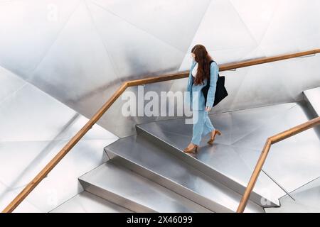 Von oben gesehen, die gesichtslose, ferne Frau mit langen Haaren in eleganter Kleidung, mit einer Handtasche, die im Flur eines hellen, geräumigen Gebäudes nach oben geht Stockfoto