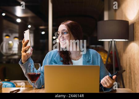 Lächelnde, intelligente junge Frau in einer Brille, die Daten auf dem Smartphone liest und analysiert, während sie während der Arbeit im Restaurant mit Gadgets und Rotwein am Tisch sitzt Stockfoto