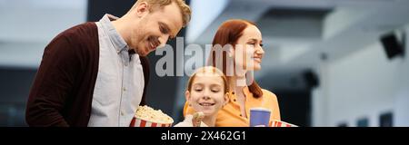 Ein Mann steht neben einer Frau und einem Kind, beide lächeln, während sie eine Kiste Popcorn in einem Kino hält. Stockfoto